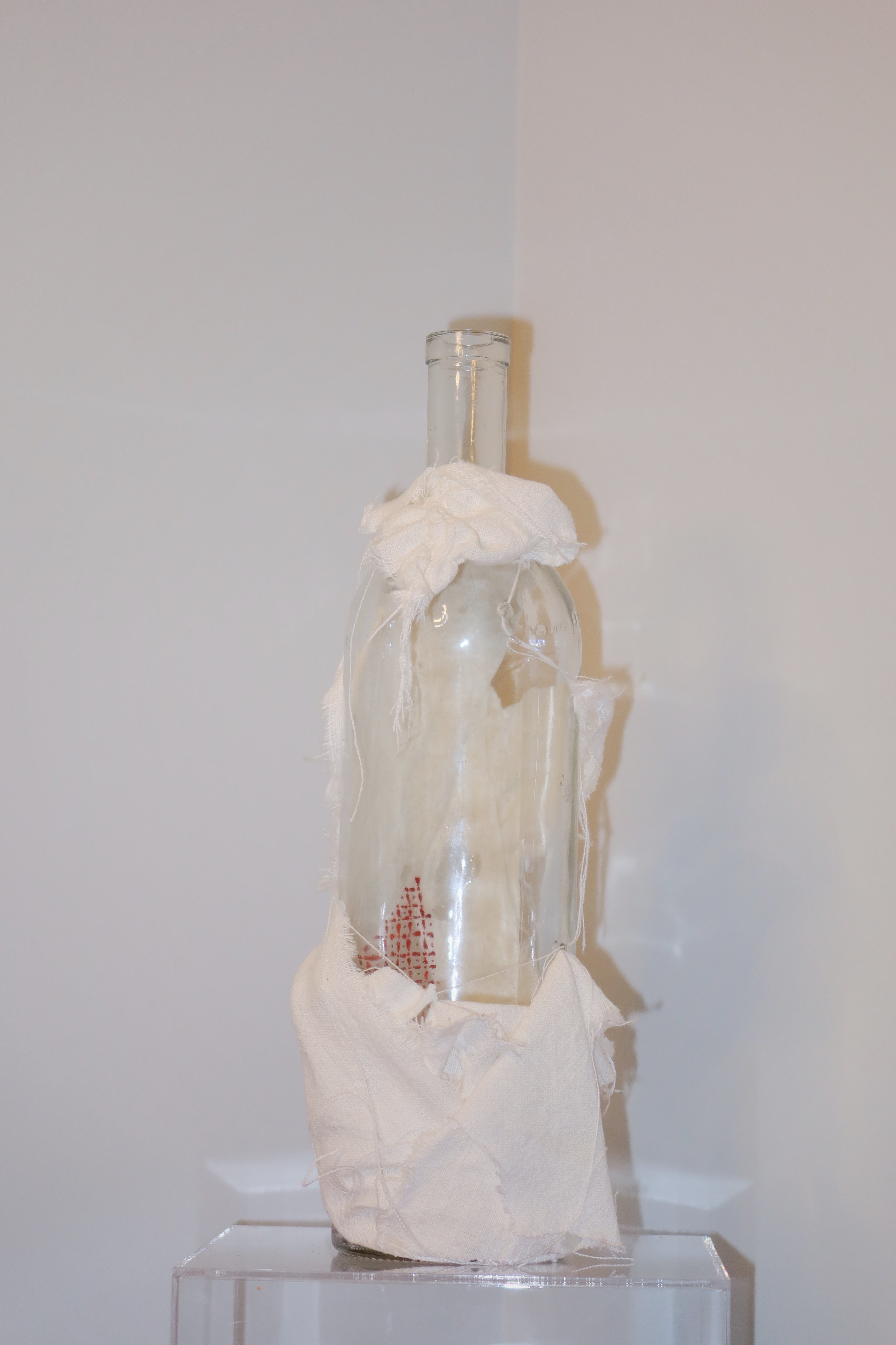 Glass bottle with decorative white linen on a clear stand against a plain background