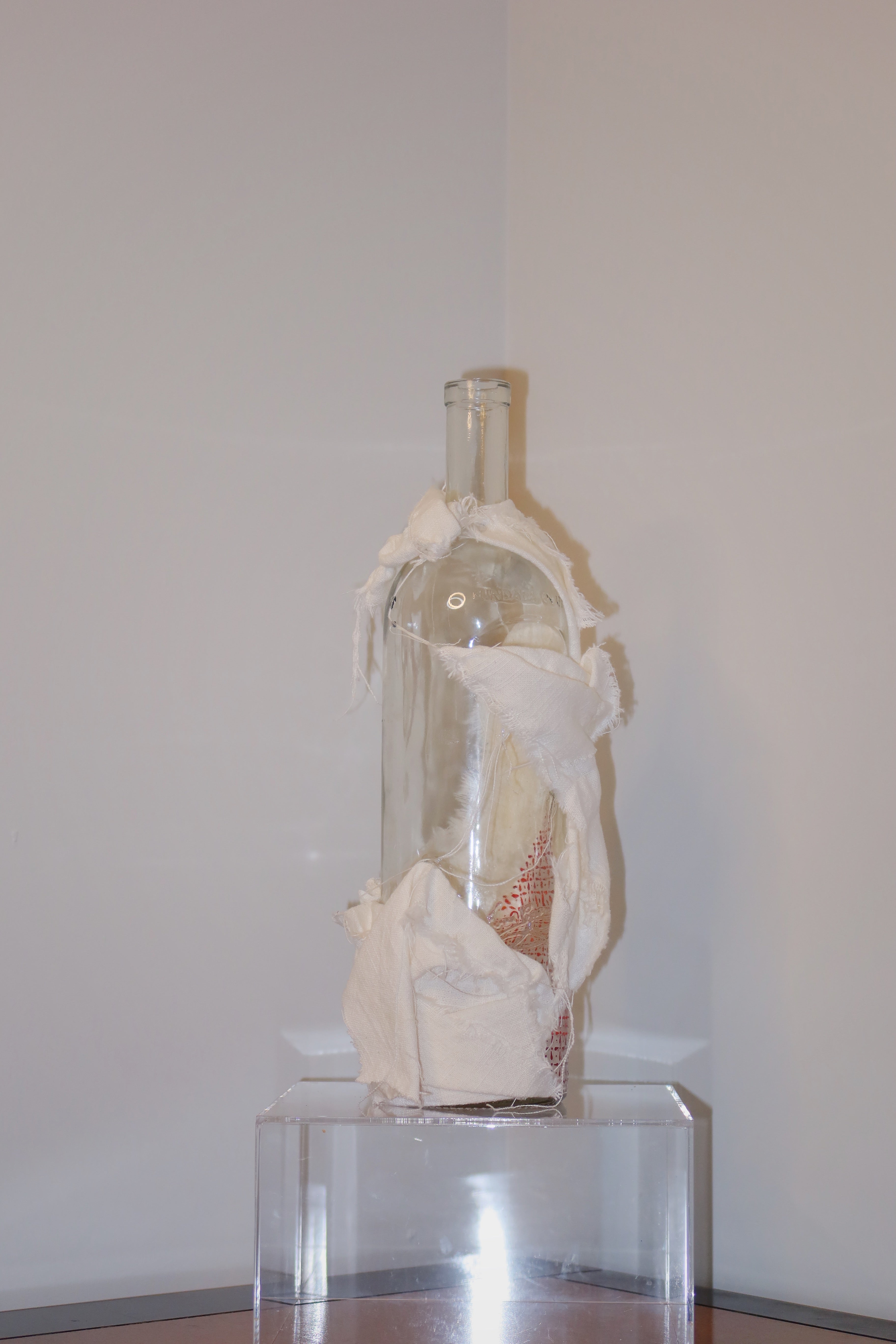 Glass bottle wrapped in white linen on a clear stand against a plain wall