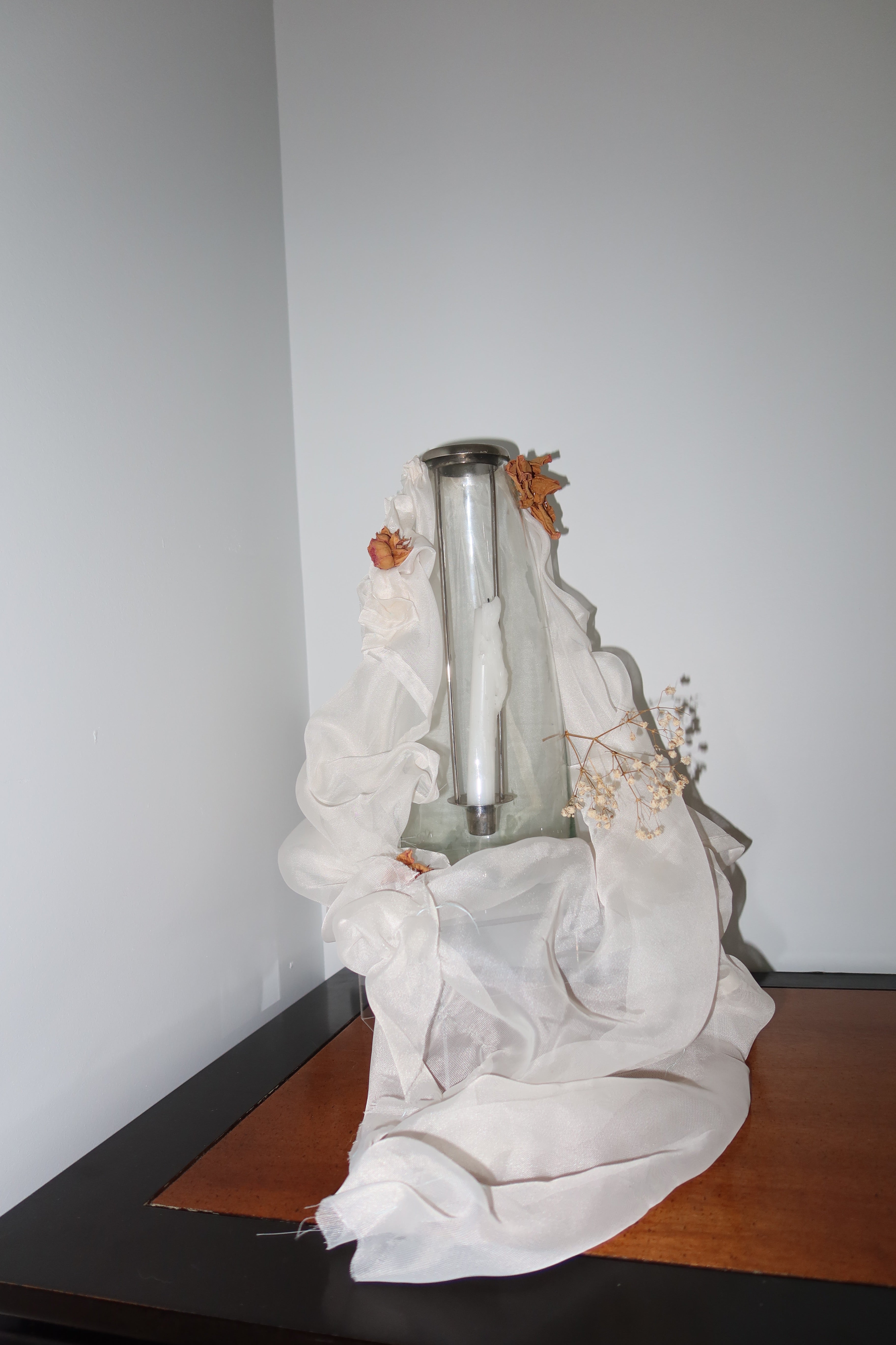 Exposed clear candle vase with white mesh fabric draped over it, little roses and baby breath embellishing the light weight cream fabric, standing on a lack and wooden table against a white bakground