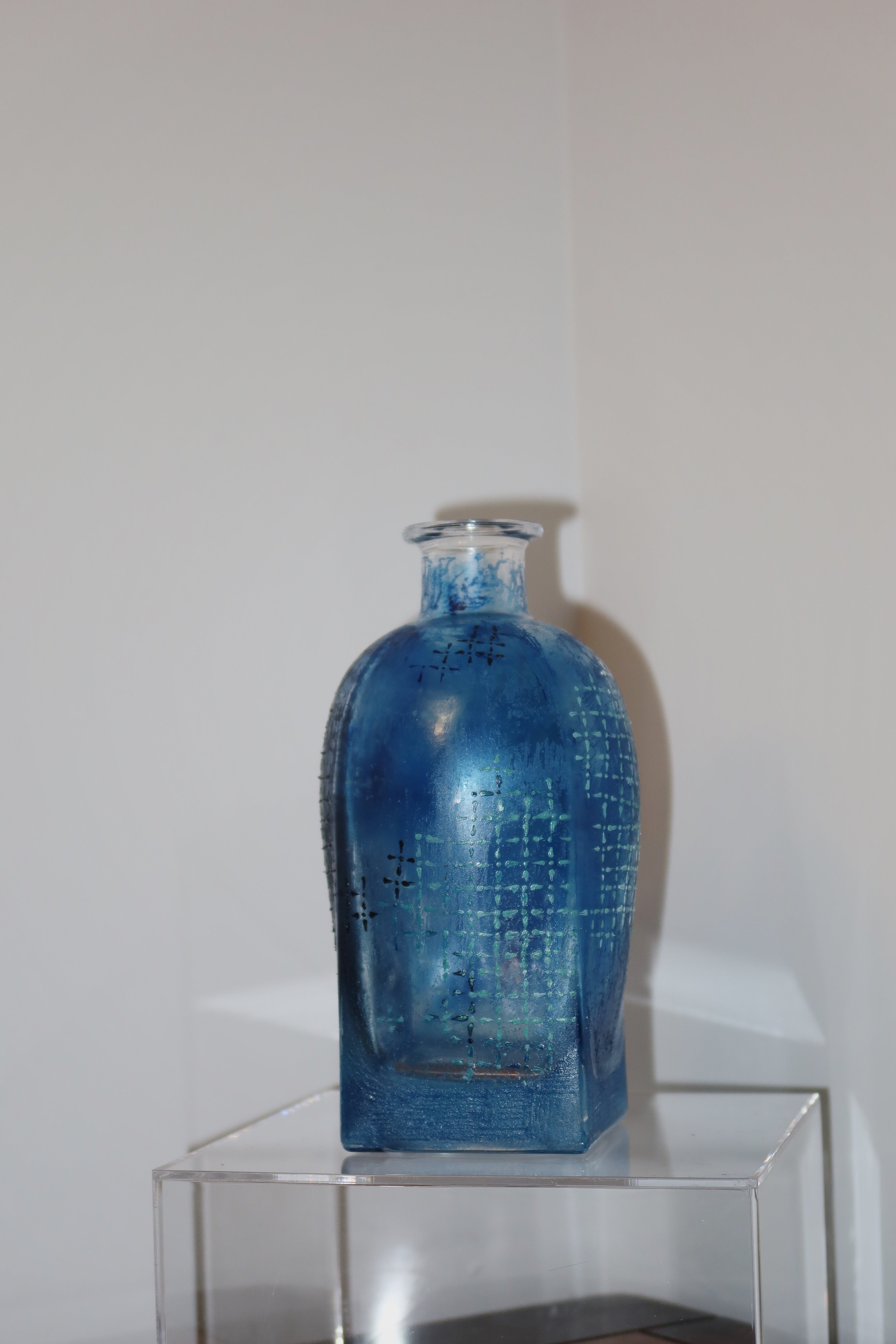 Blue glass bottle with geometric pattern on a clear stand against a white wall