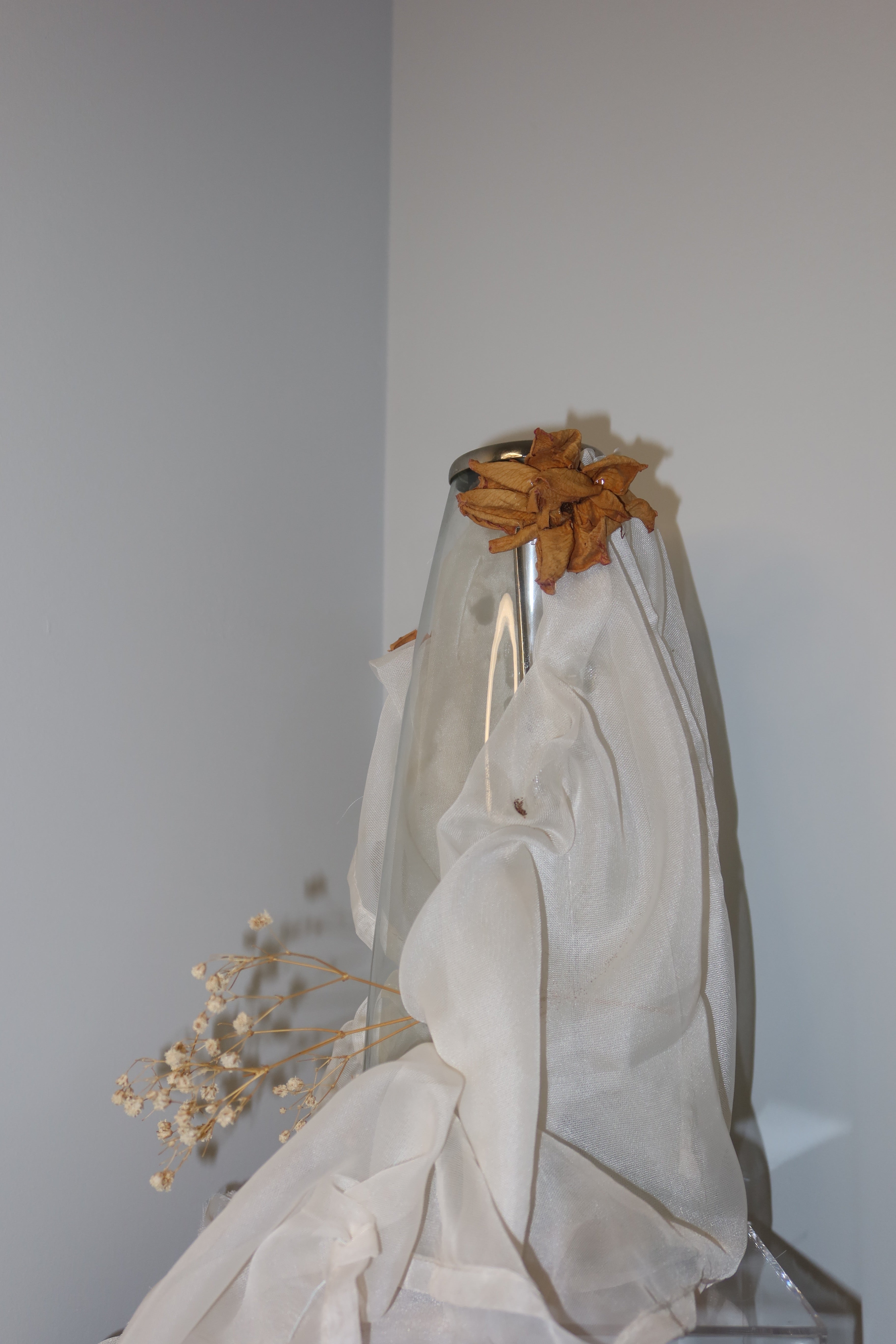 close up of clear vase from the side with white mesh fabric draped over it, little roses and baby breath embellishing the light weight cream fabric, standing on a wooden surface against a white background