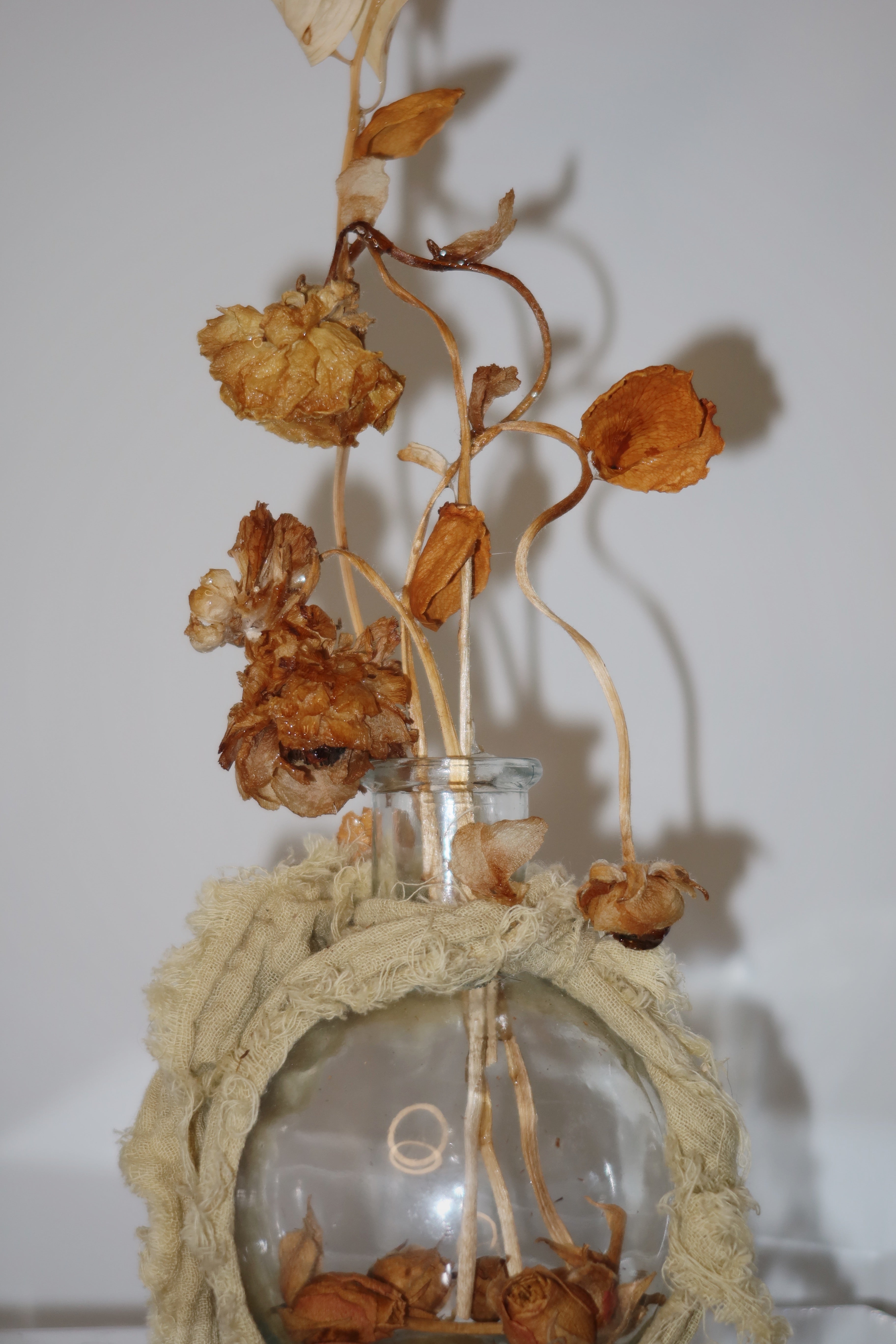 Glass vase wrapped in a sage green linen, with dried flowers and leaves reaching high up. standing on a clear surface against a white background