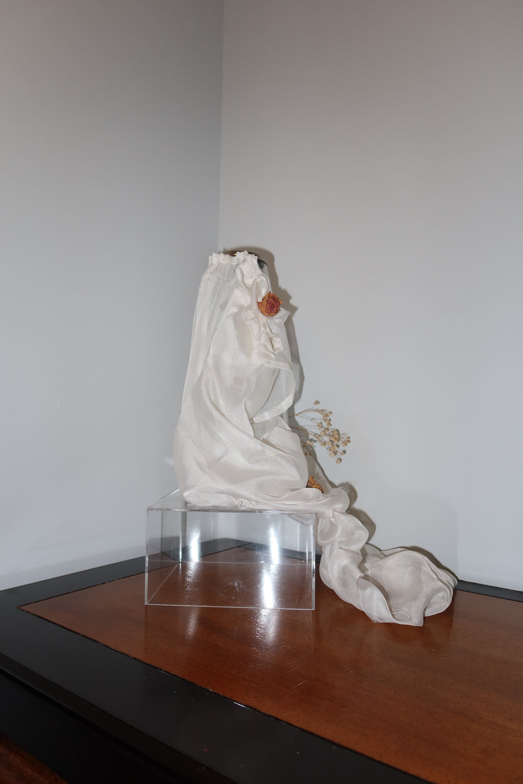 of clear vase with white mesh fabric draped over it, little roses and baby breath embellishing the light weight cream fabric, standing on a wooden surface against a white background
