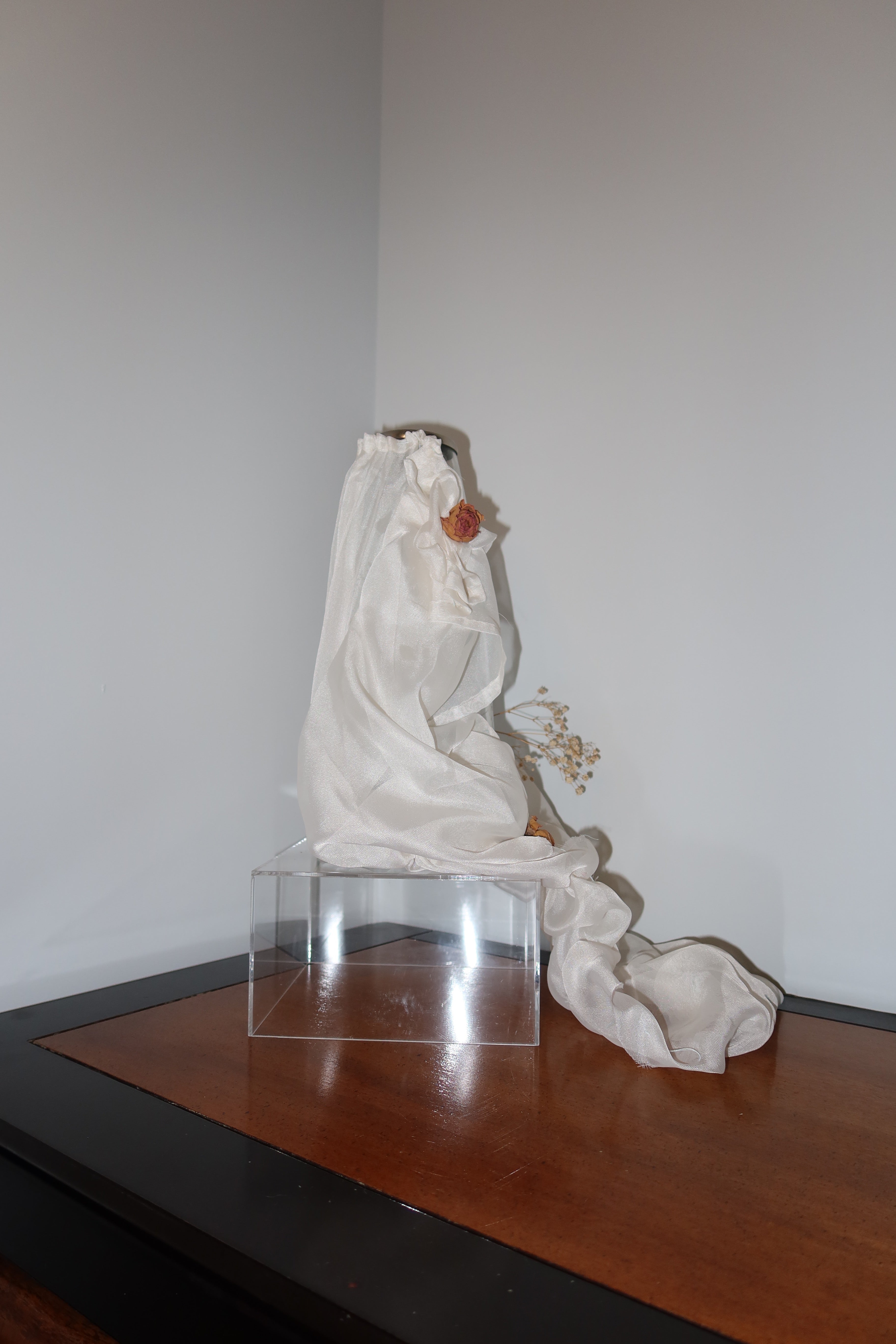 of clear vase with white mesh fabric draped over it, little roses and baby breath embellishing the light weight cream fabric, standing on a wooden surface against a white background