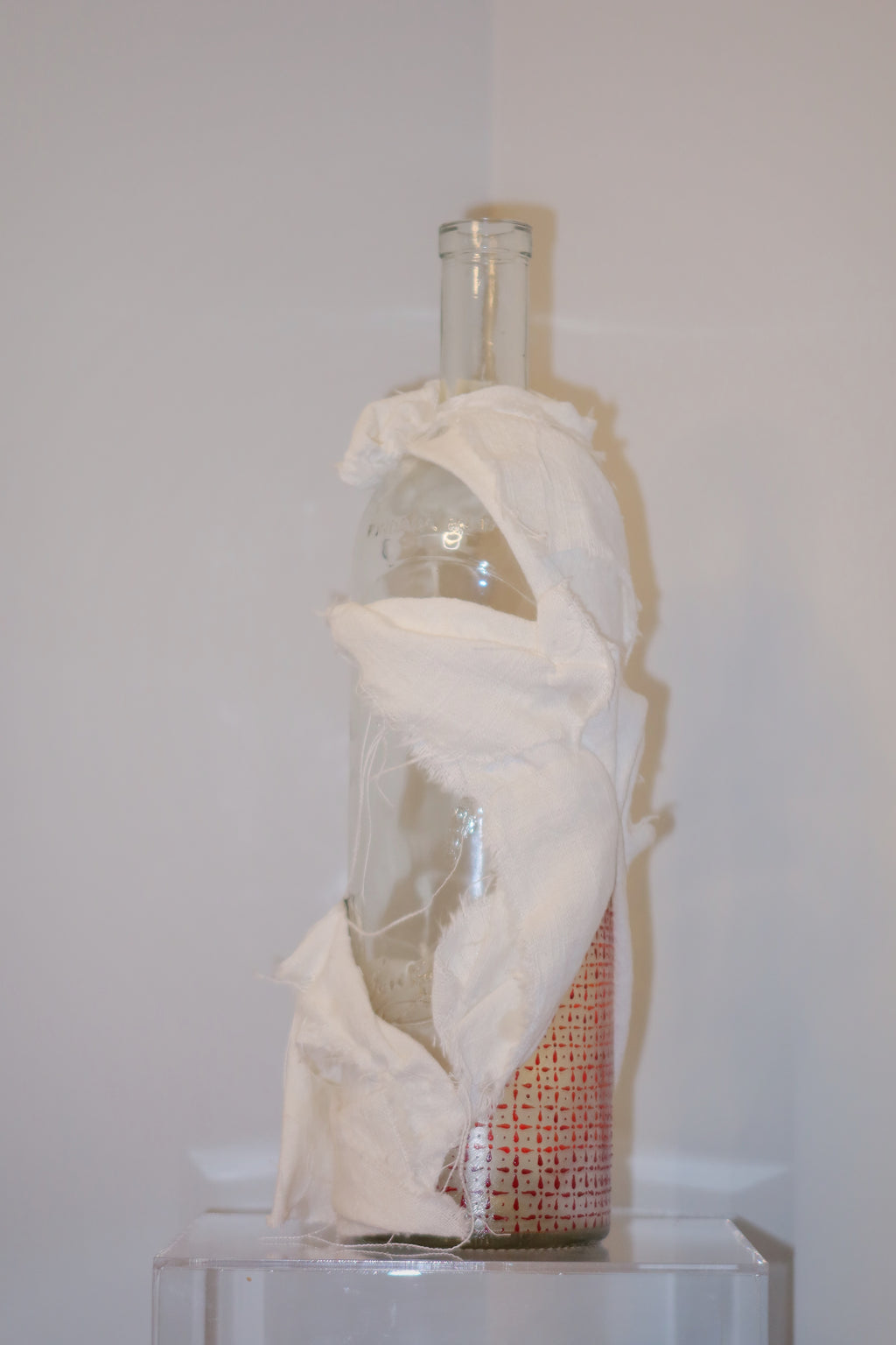 Glass bottle wrapped in white cloth on a white pedestal against a white wall