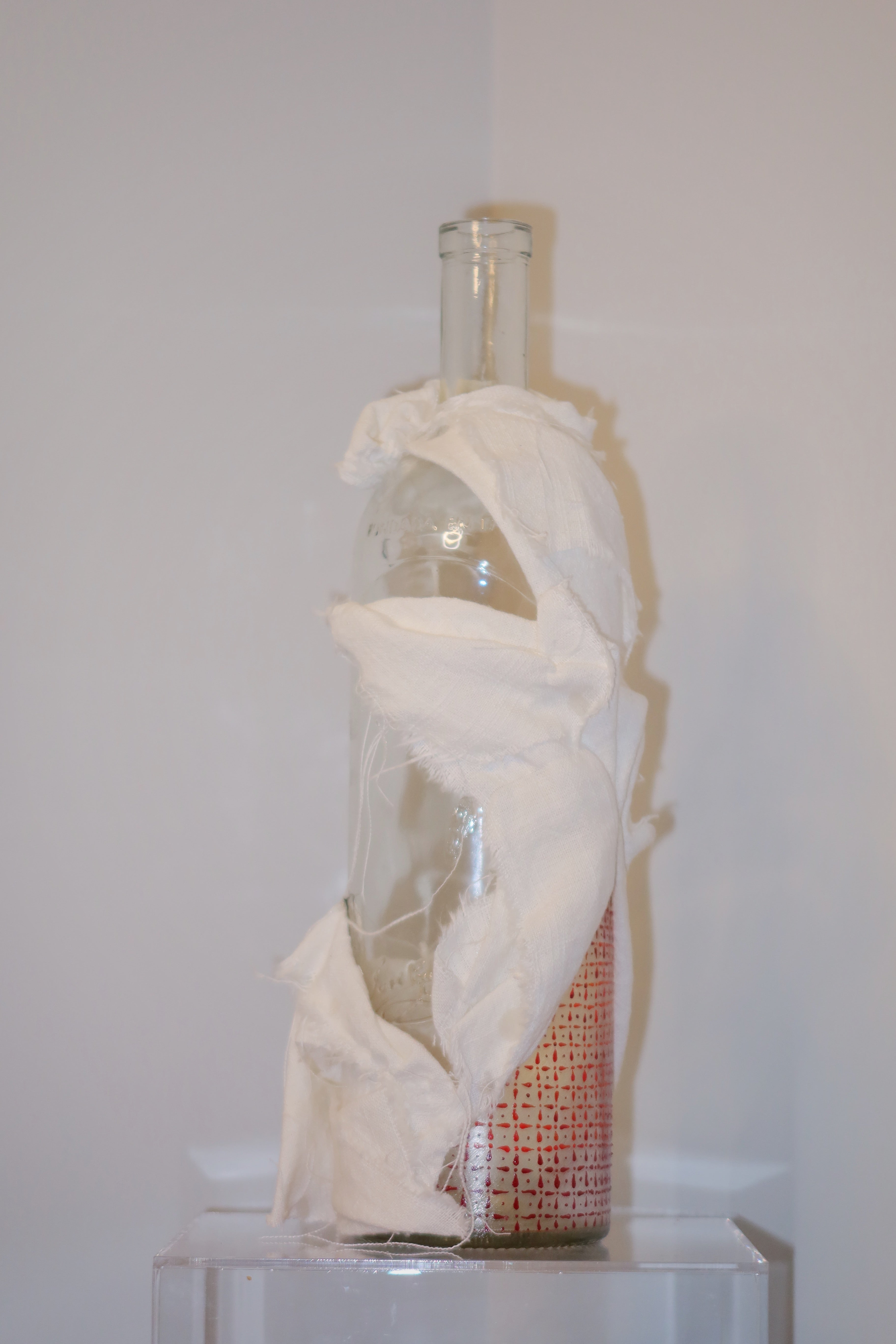Glass bottle wrapped in white cloth on a white pedestal against a white wall