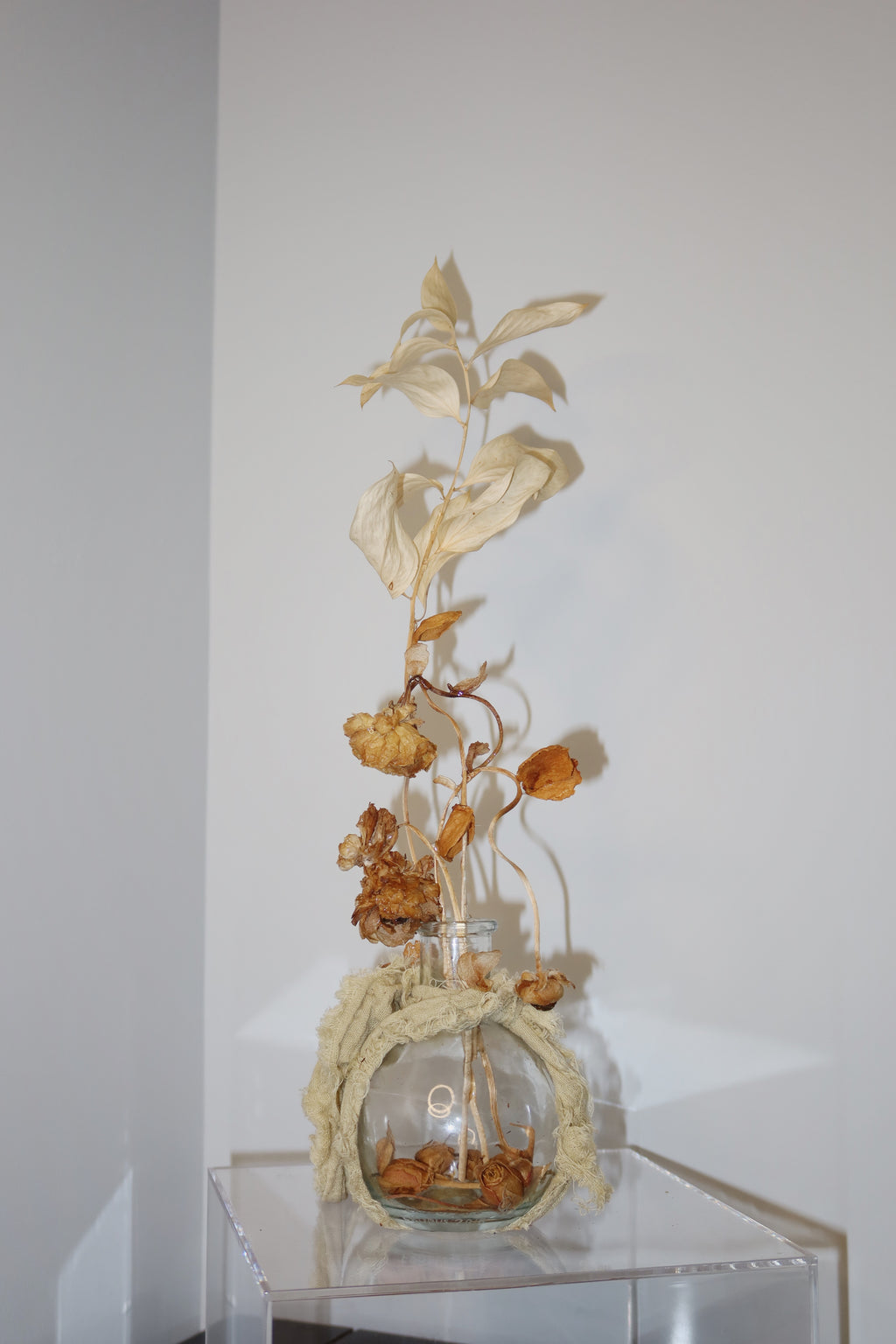 Glass vase wrapped in a sage green linen, with dried flowers and leaves reaching high up. standing on a clear surface against a white background