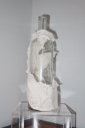 Glass bottle filled with white material on a clear stand against a plain wall