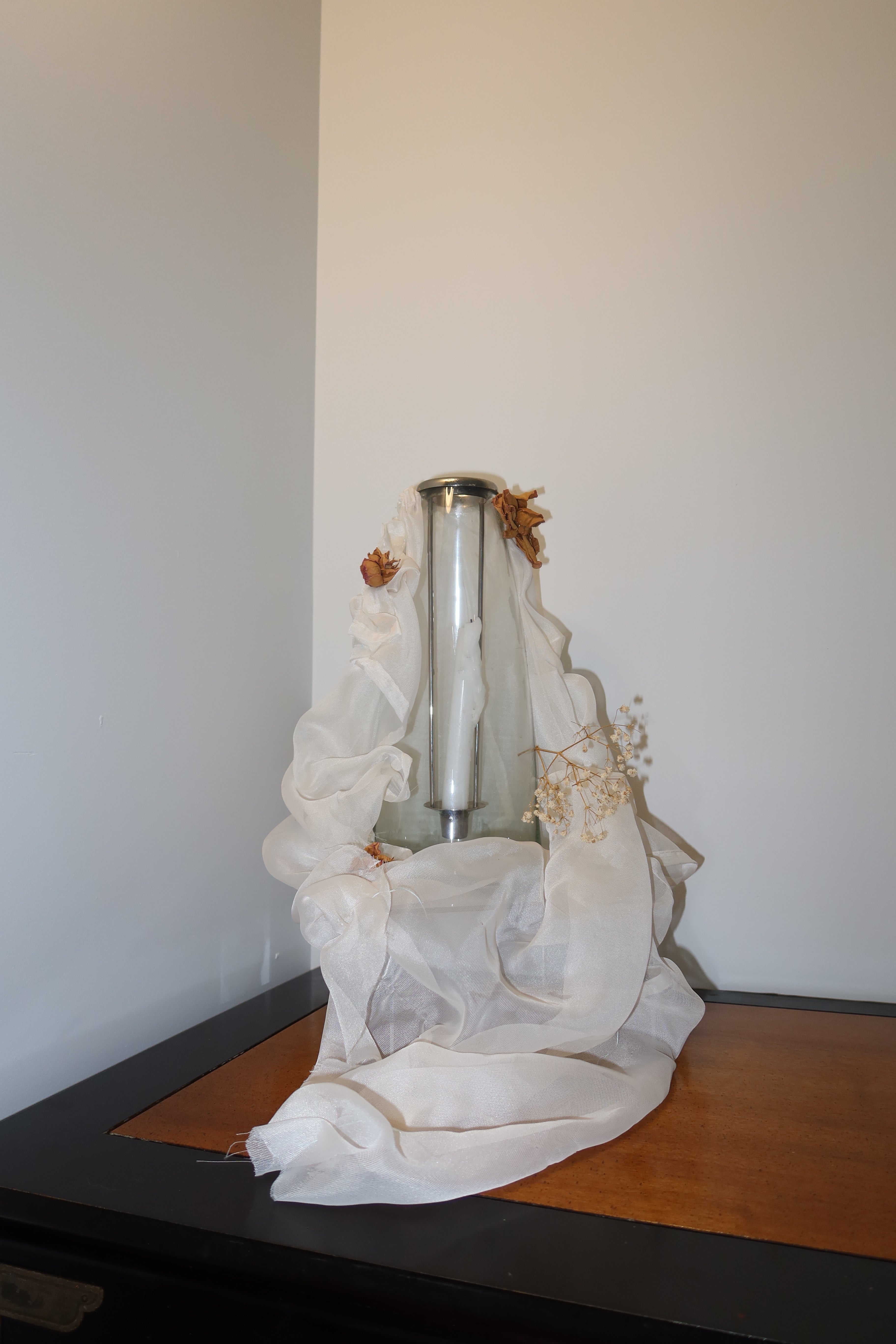close up of clear candle vase with white mesh fabric draped over it, little roses and baby breath embellishing the light weight cream fabric, standing on a wooden surface against a dark red background