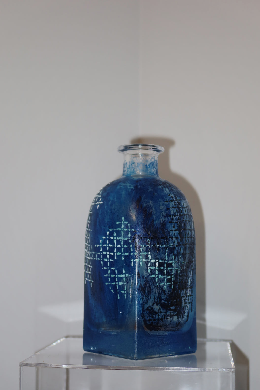 Blue glass bottle with geometric pattern on a clear stand against a white background