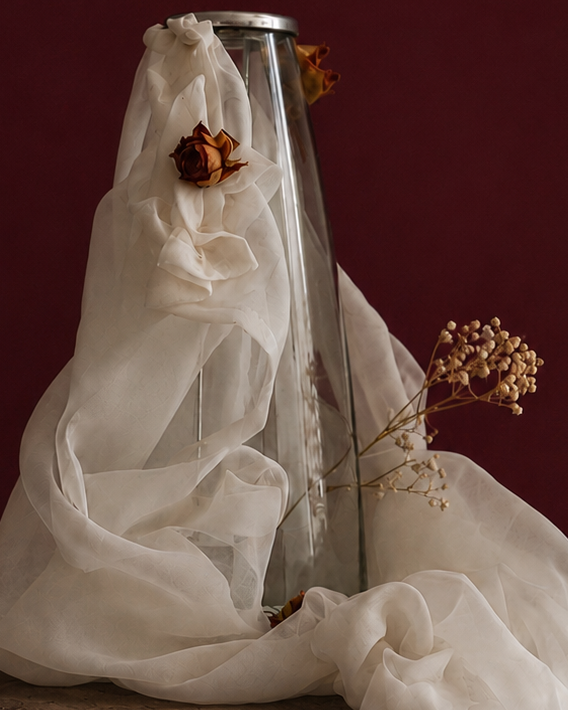 close up of clear candle vase with white mesh fabric draped over it, little roses and baby breath embellishing the light weight cream fabric, standing on a wooden surface against a dark red background