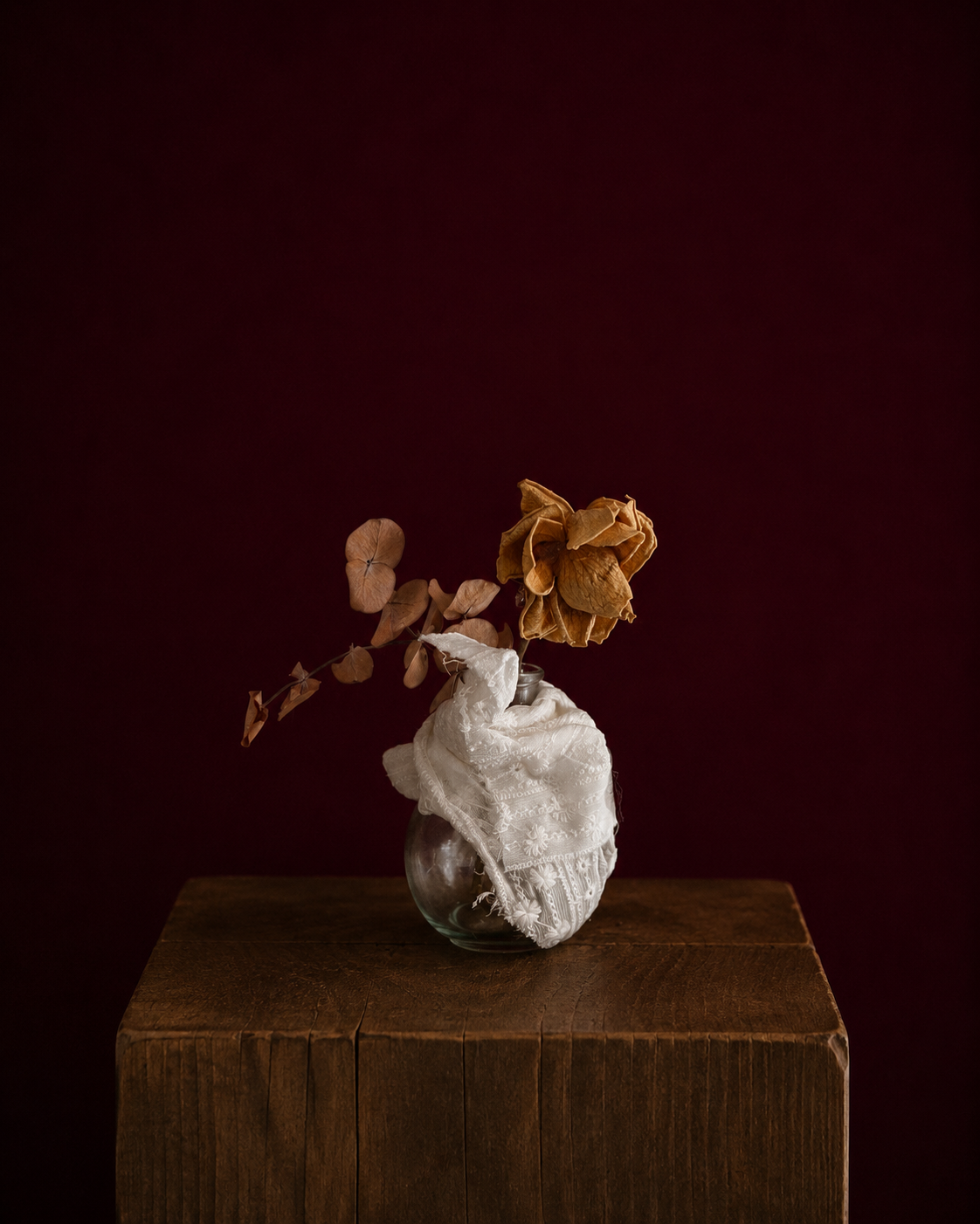 round vase wrapped in eyelet fabric, holding a yellow rose and eucalyptus leaf.