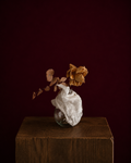round vase wrapped in eyelet fabric, holding a yellow rose and eucalyptus leaf.