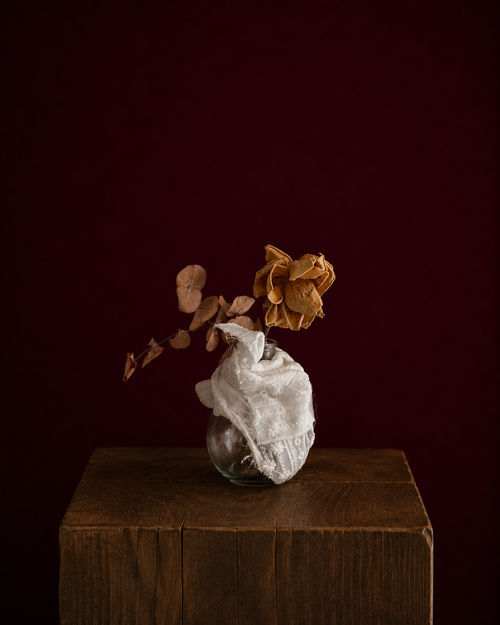 round vase wrapped in eyelet fabric, holding a yellow rose and eucalyptus leaf.