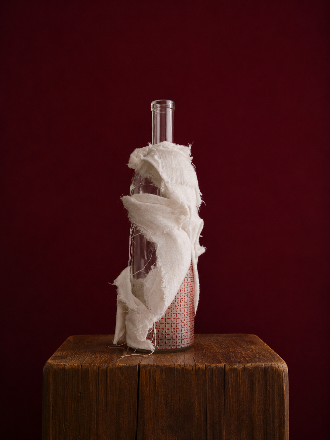 Glass bottle with white linen
, handpainted pattern, on a wooden block against a dark red background