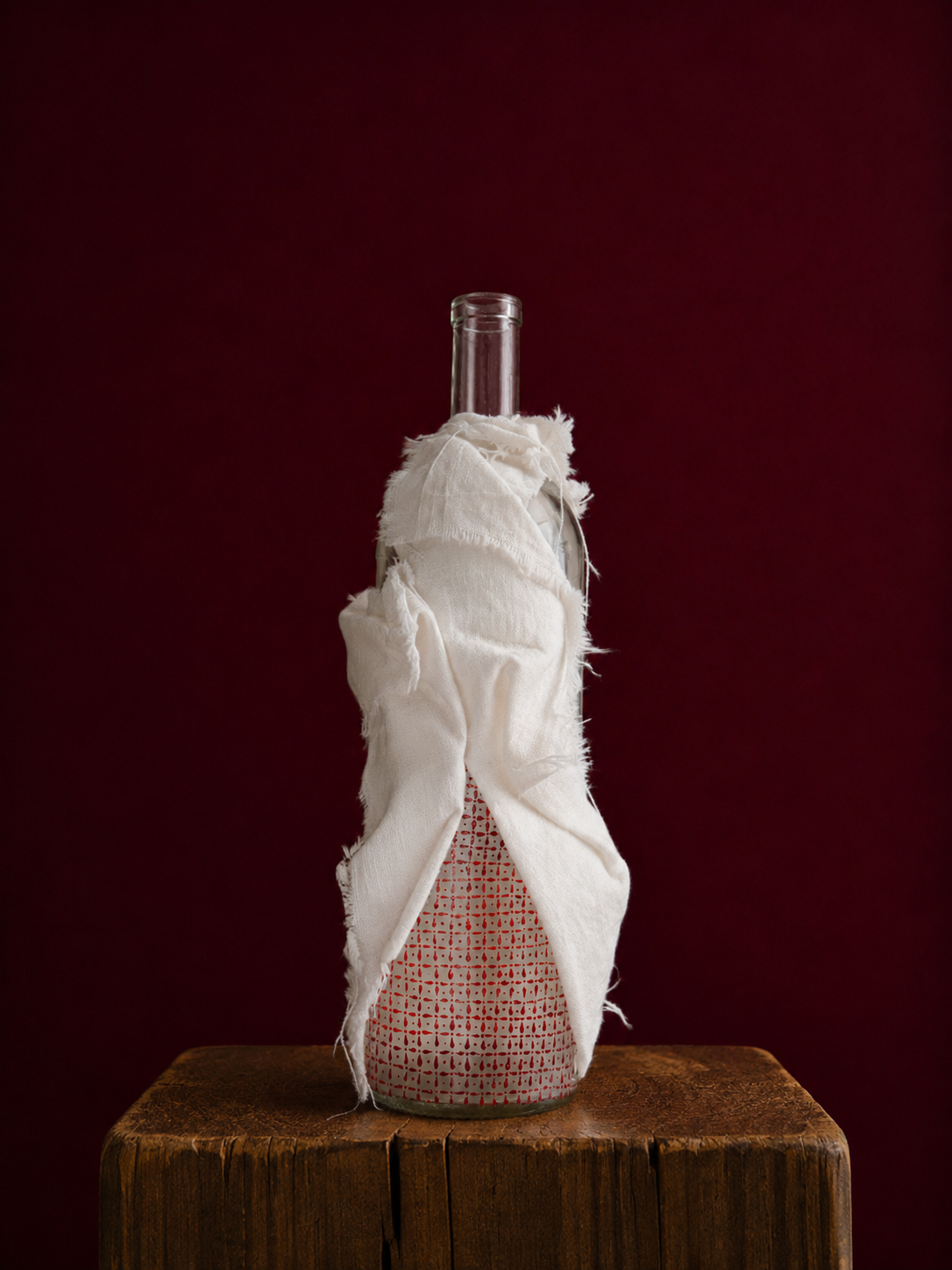 Decorative bottle wrapped in white linen on a wooden surface with a dark red background