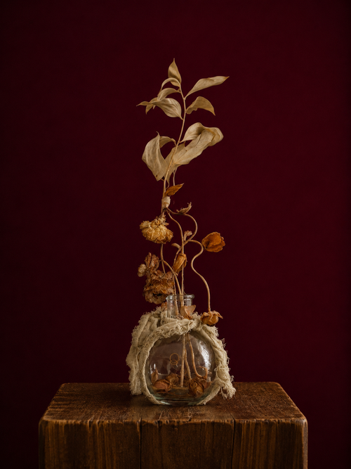 Glass vase wrapped in a sage green linen, with dried flowers and leaves reaching high up. standing on a wooden surface against a dark red background