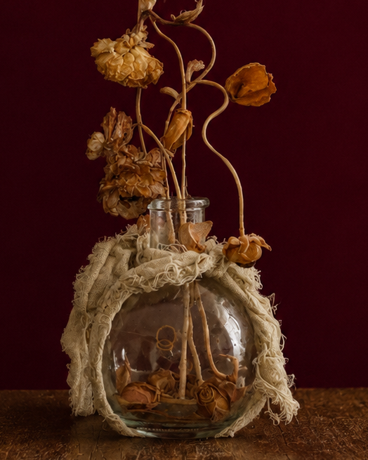 Up-close glass vase wrapped in a sage green linen, with dried flowers and leaves reaching high up. standing on a wooden surface against a dark red background