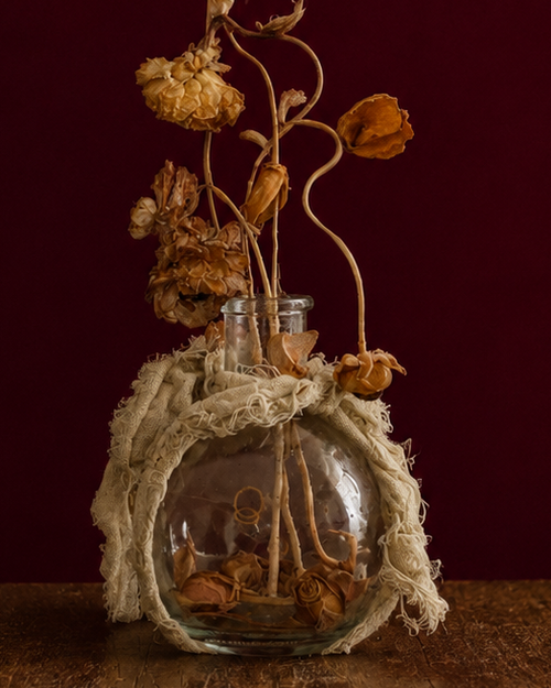 Up-close glass vase wrapped in a sage green linen, with dried flowers and leaves reaching high up. standing on a wooden surface against a dark red background
