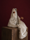 Clear candle vase with white mesh fabric draped over it, little roses embellishing the light weight cream fabric, standing on a wooden surface against a dark red background
