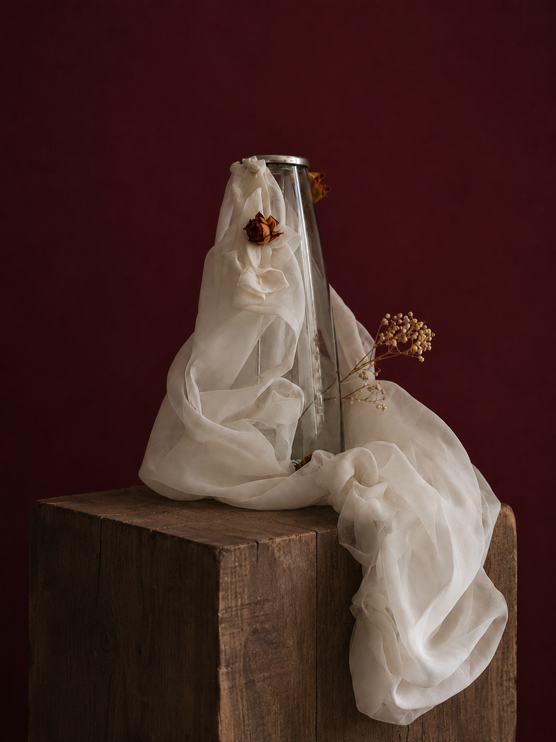 Clear candle vase with white mesh fabric draped over it, little roses embellishing the light weight cream fabric, standing on a wooden surface against a dark red background