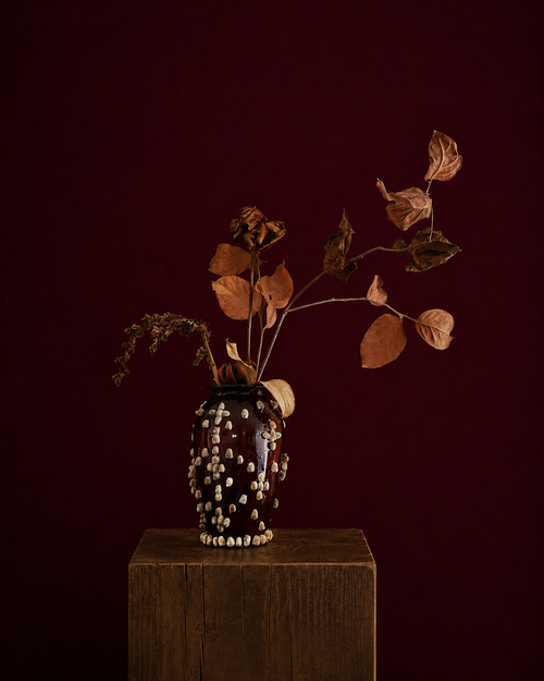 Decorative vase with dried leaves and glued on fluffy seeds, sitting on a wooden pedestal against a dark red background