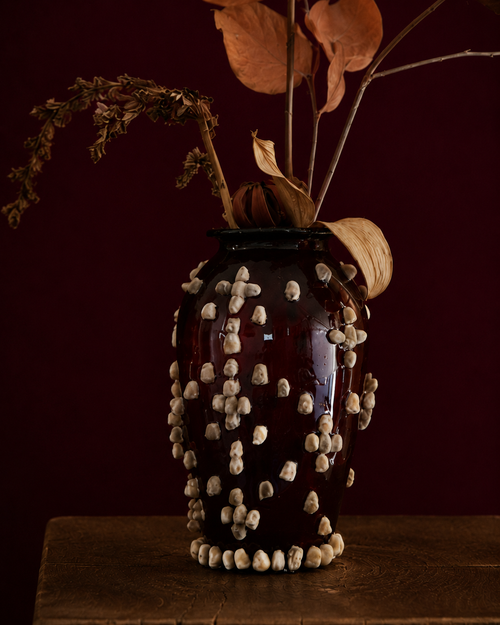 Decorative vase with dried leaves and glued on fluffy seeds, sitting on a wooden pedestal against a dark red background