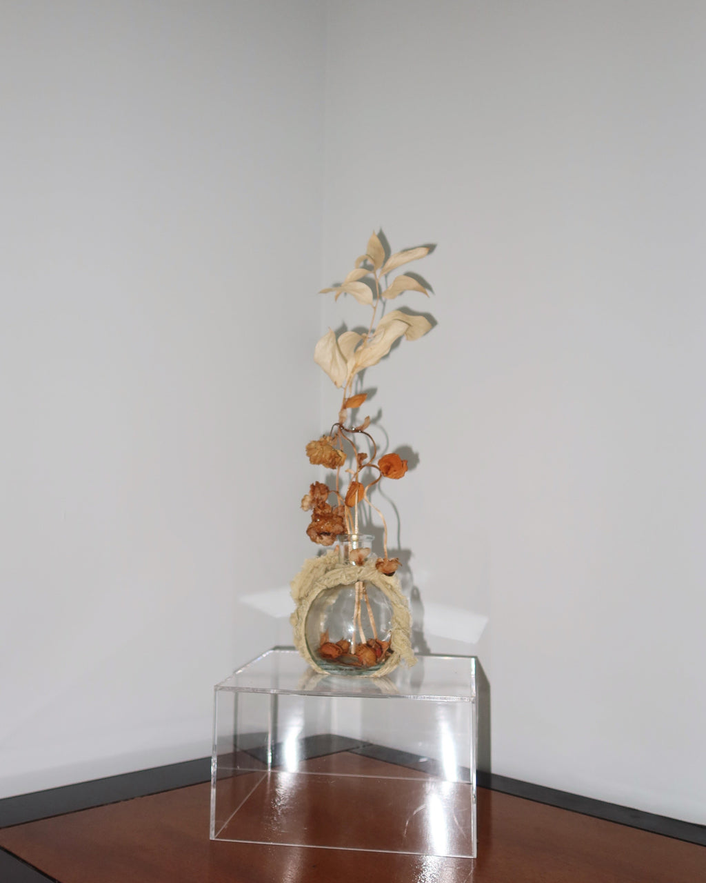 Glass vase wrapped in a sage green linen, with dried flowers and leaves reaching high up. standing on a clear surface against a white background