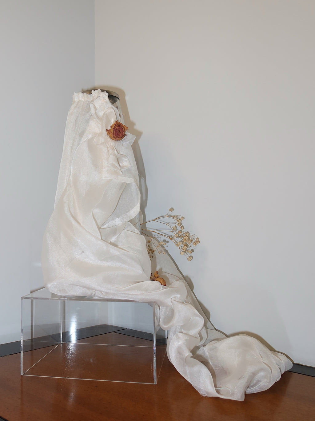side of cancel vase with white mesh fabric draped over it, little roses and baby breath embellishing the light weight cream fabric, standing on a wooden surface against a dark red background
