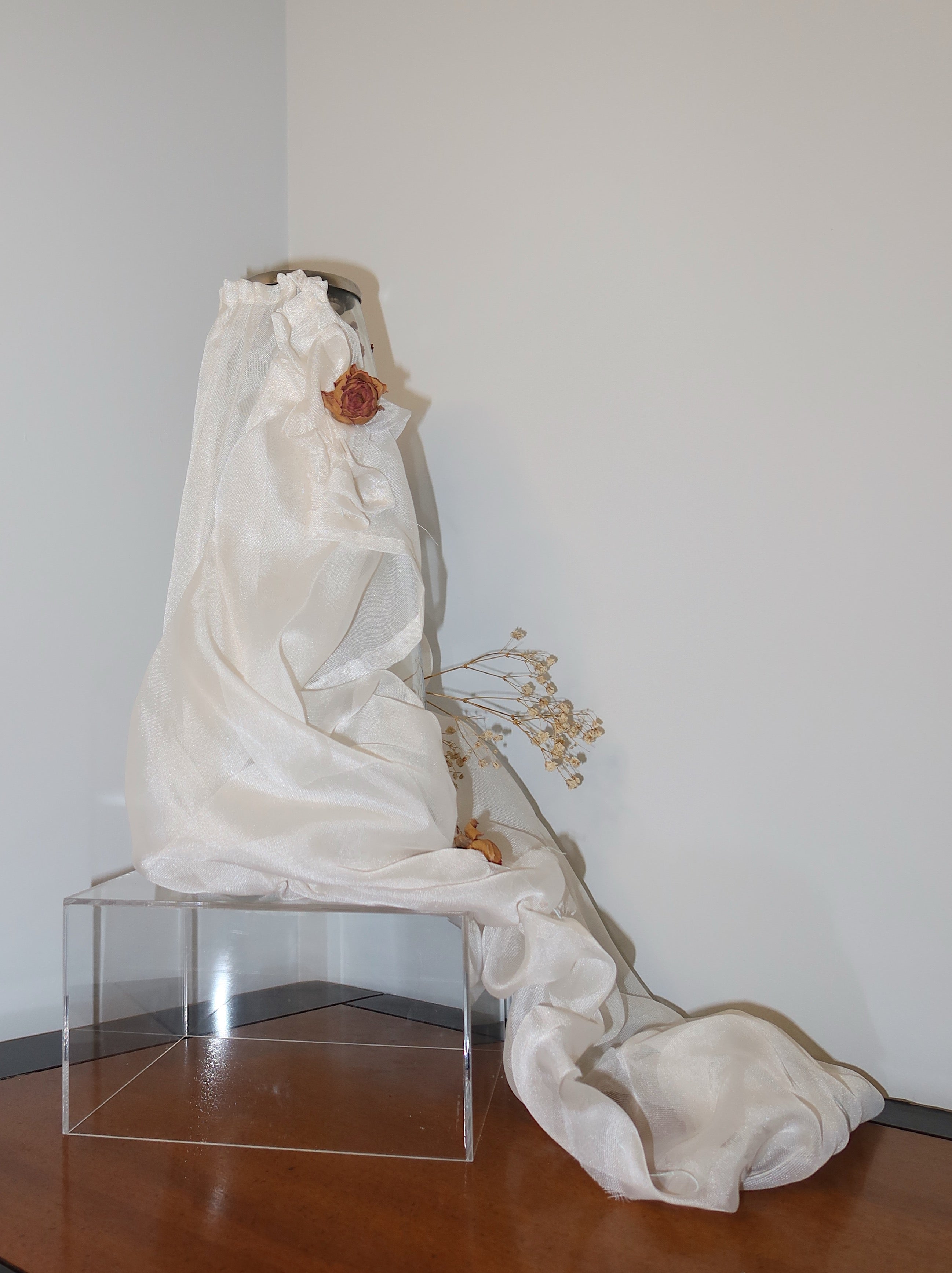side of cancel vase with white mesh fabric draped over it, little roses and baby breath embellishing the light weight cream fabric, standing on a wooden surface against a dark red background