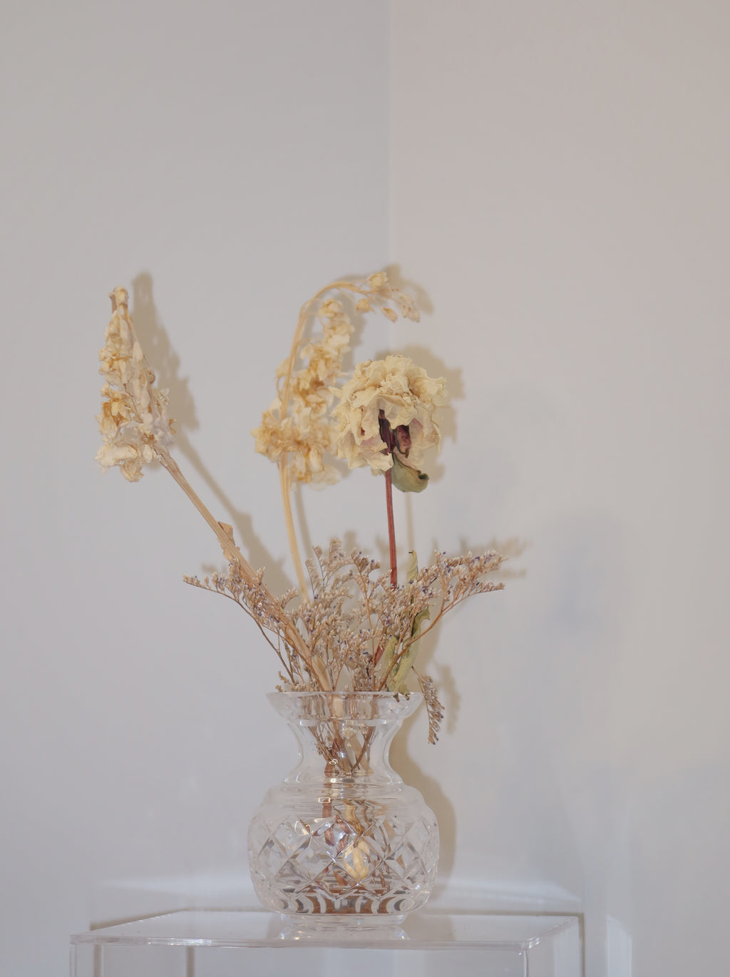 crystal vase with dried peony and other flowers on a clear surface against a white background.