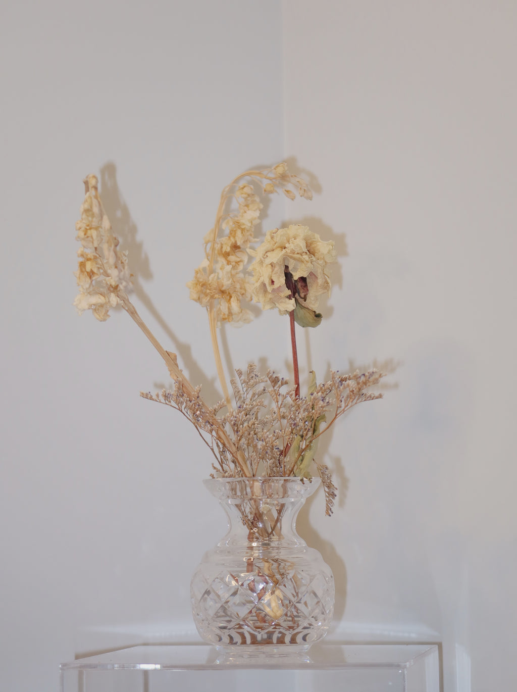 crystal vase with dried peony and other flowers on a clear surface against a white background.