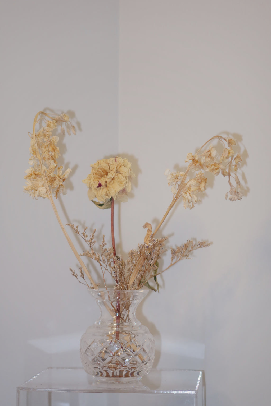 crystal vase with dried peony and other flowers on a clear surface against a white background.