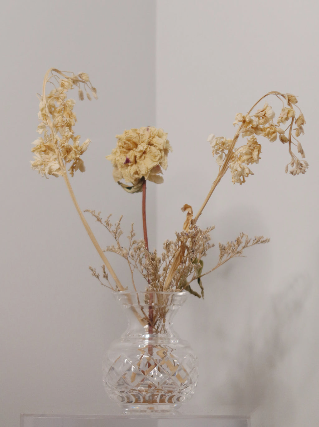 crystal vase with dried peony and other flowers on a clear surface against a white background.