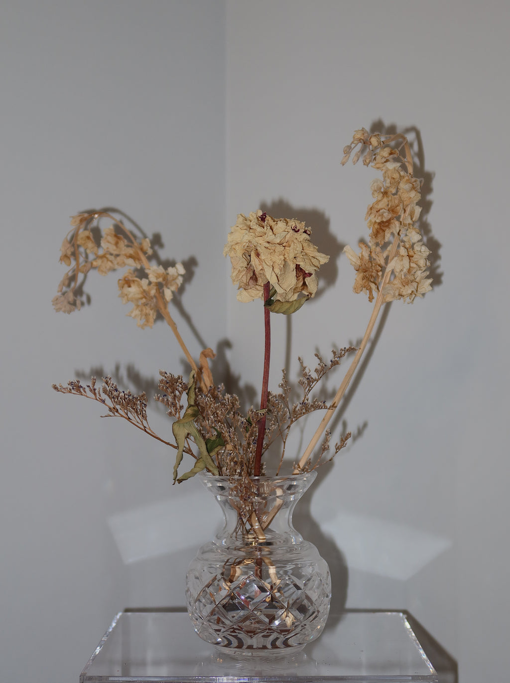 crystal vase with dried peony and other flowers on a clear surface against a white background.