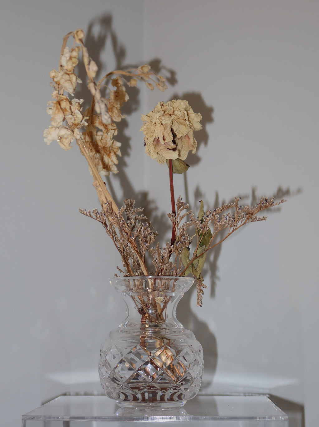 crystal vase with dried peony and other flowers on a clear surface against a white background.