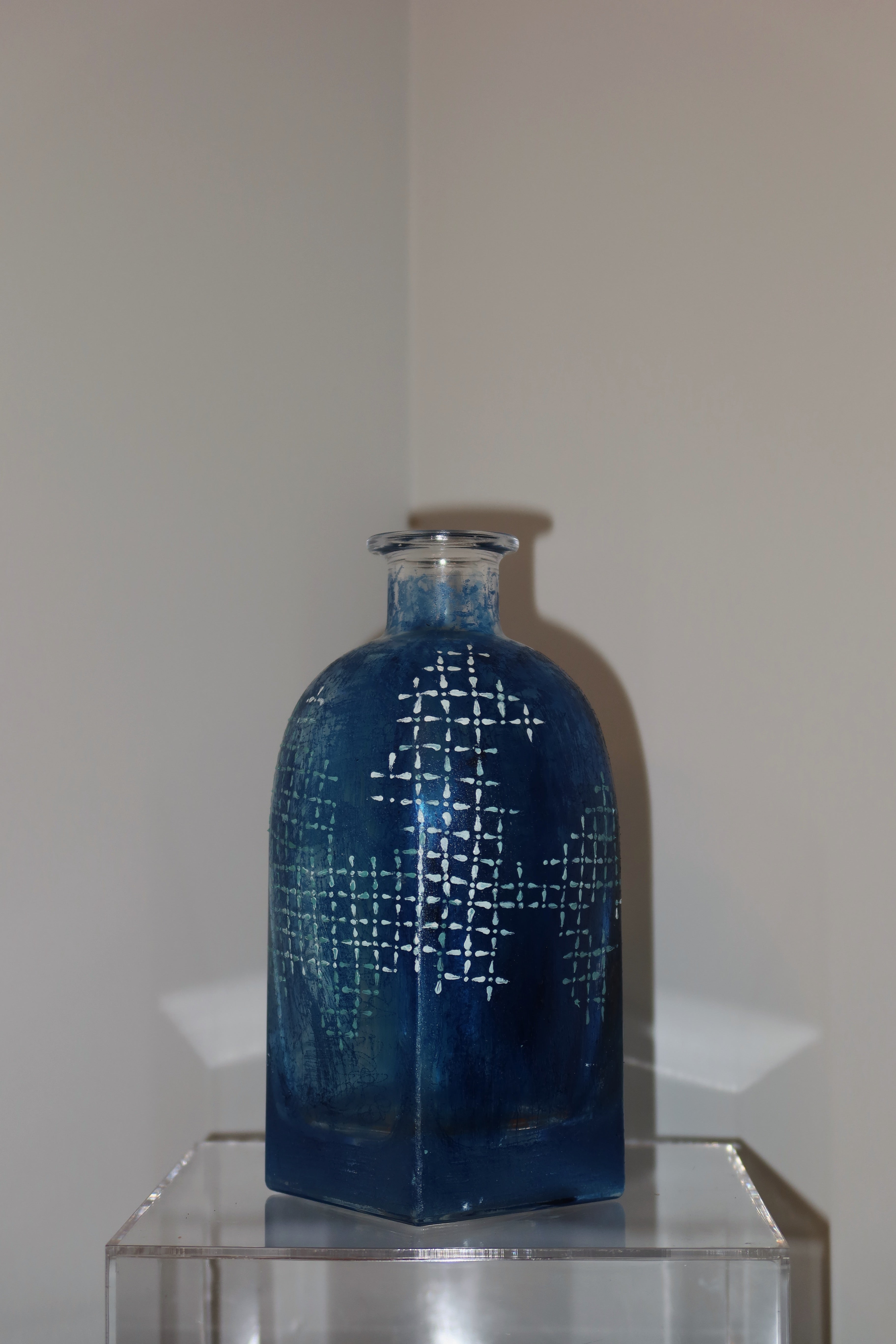 Blue glass bottle with geometric pattern on a clear stand against a plain wall