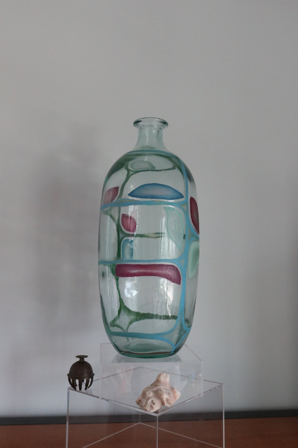 A tall, narrow glass vase with abstract blue, green, and magenta patterns, displayed on a clear acrylic pedestal on a wooden table against a plain gray wall.