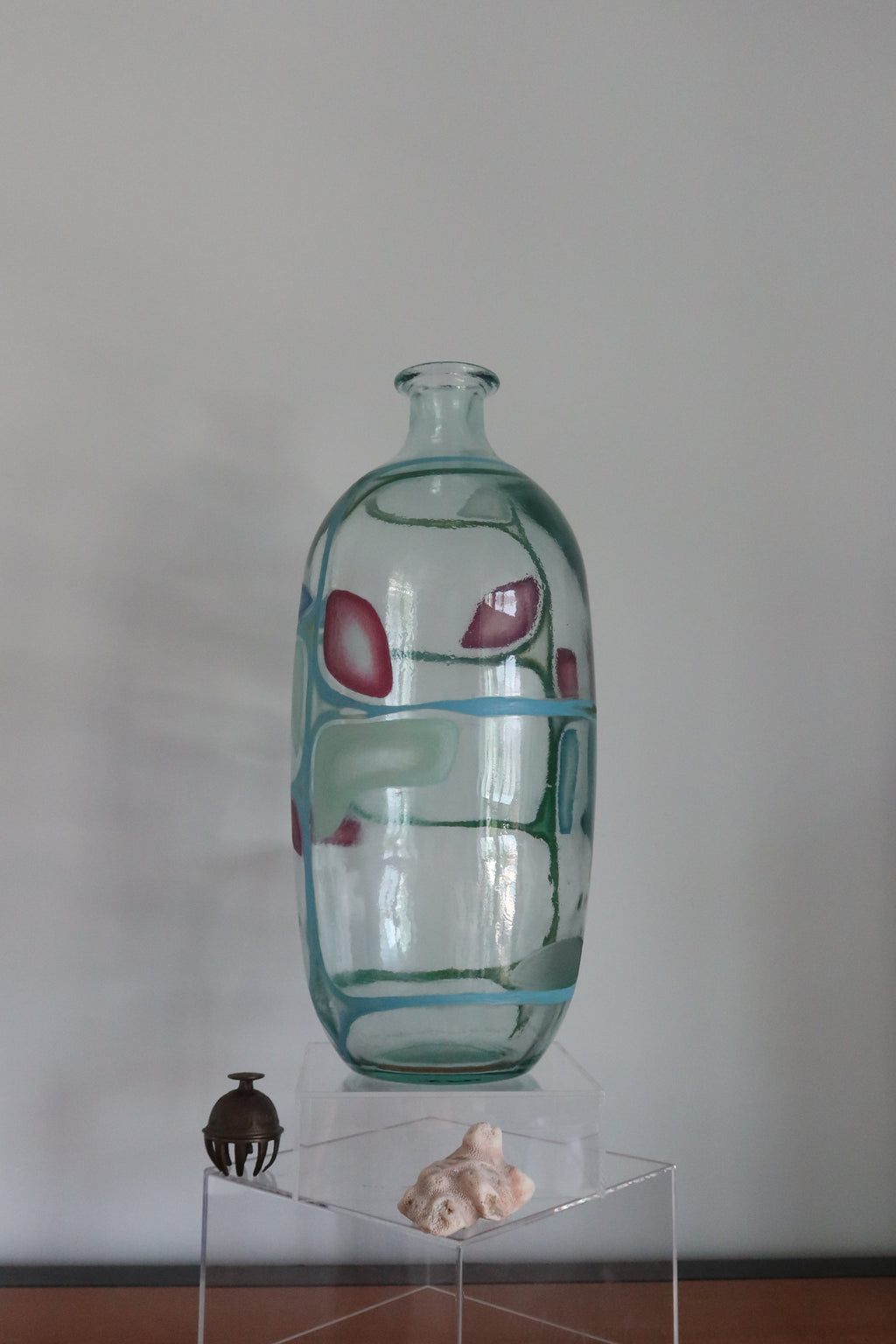 A clear glass vase with abstract red and blue painted patterns, displayed on a transparent acrylic pedestal on a wooden table against a plain gray wall.