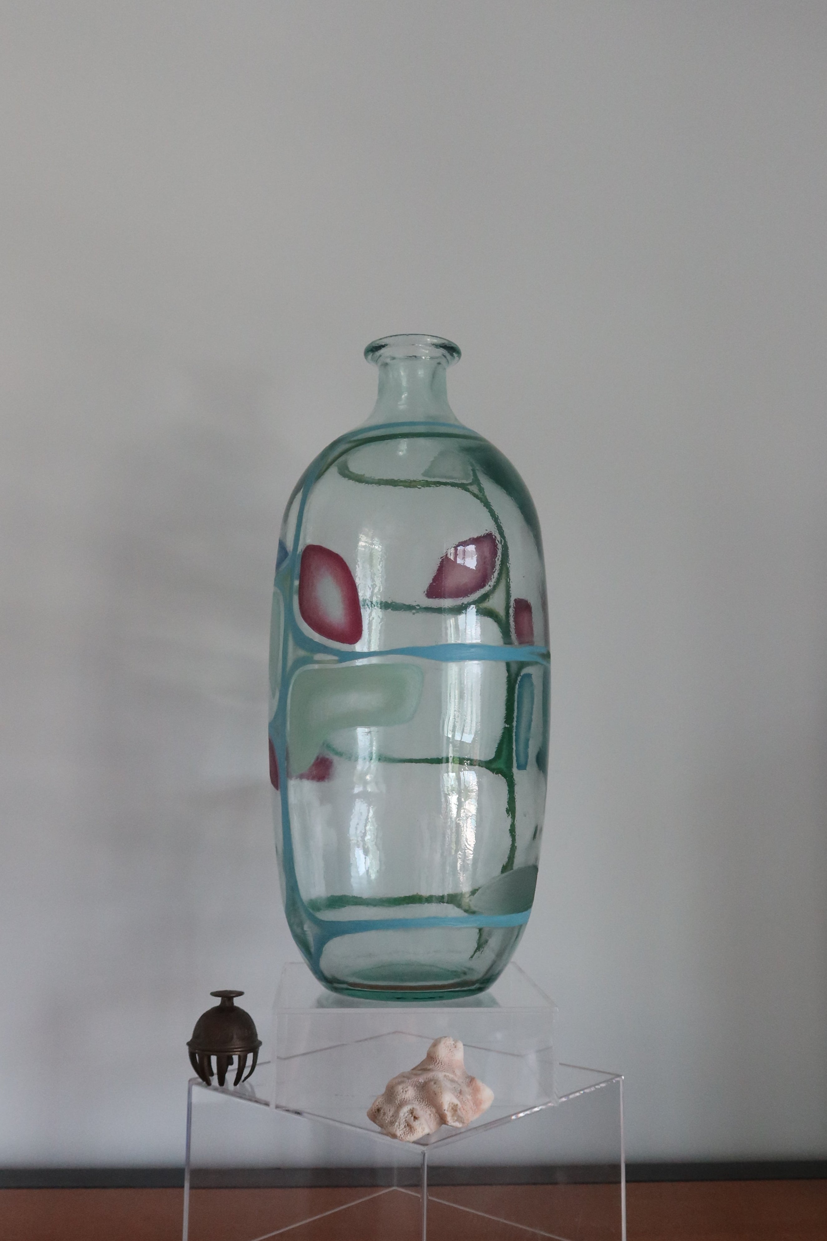 A clear glass vase with abstract red and blue painted patterns, displayed on a transparent acrylic pedestal on a wooden table against a plain gray wall.