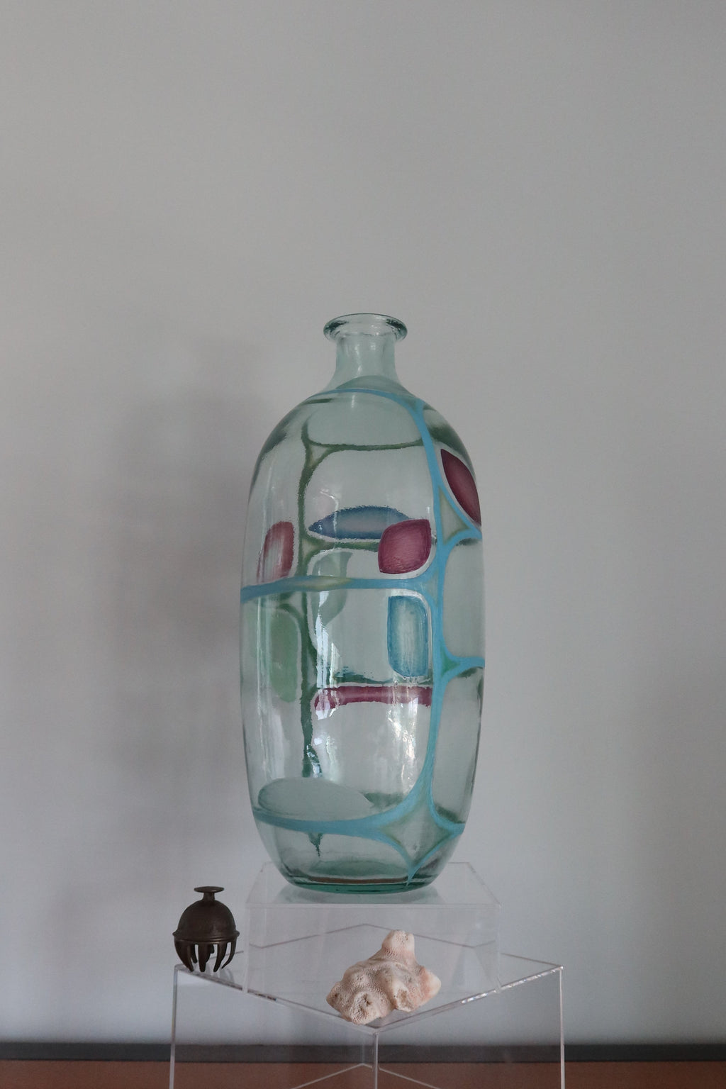 A tall, clear glass vase with a blue-green tint and pink circular accents, displayed on a clear acrylic pedestal against a plain light gray wall.