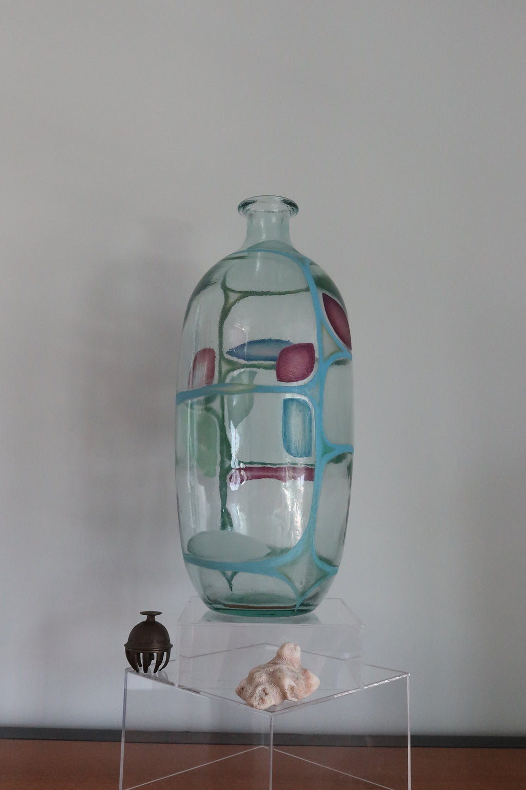 A tall, clear glass vase with a art deco patterns of light blue, purple red, and and green shapes, displayed on a clear acrylic pedestal on a wooden table against a plain gray wall.