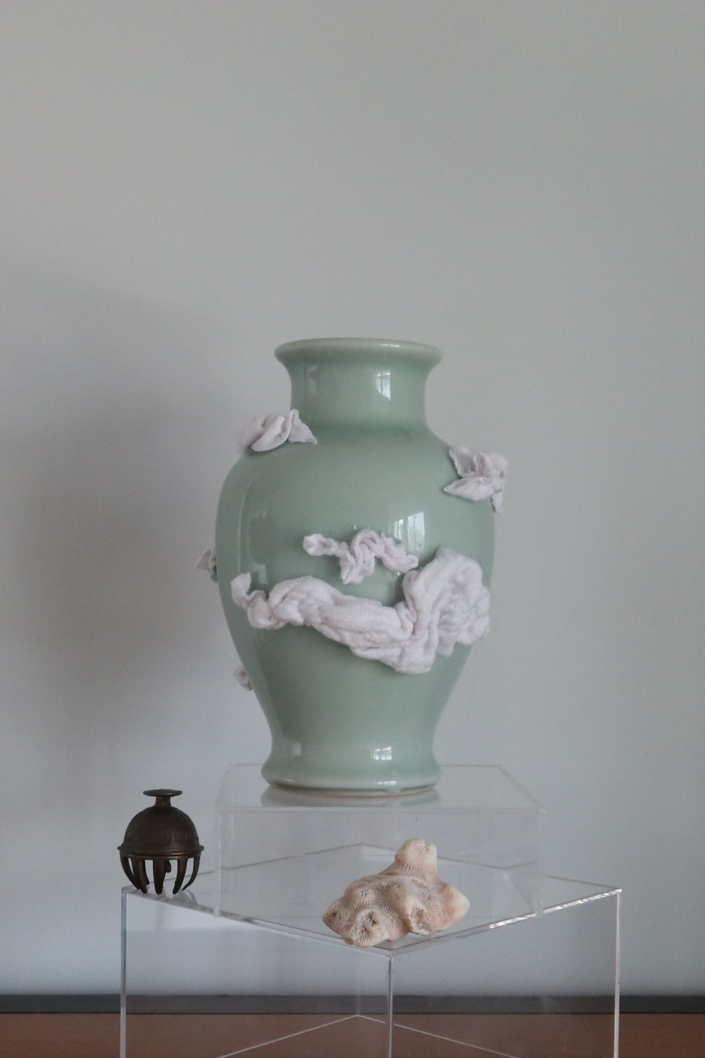 A pale green ceramic vase with white clouds made of fabric as decoration on the vase. It stands on a clear acrylic display block on a wooden table against a plain light gray wall.
