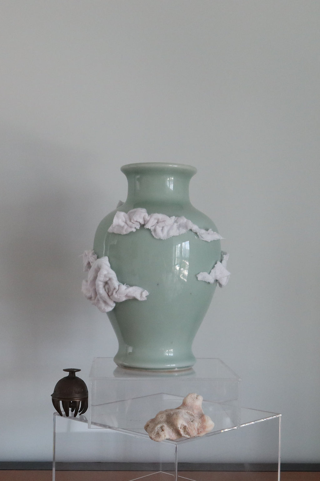 A pale celadon-green ceramic vase with white clouds made of scrap fabric, displayed on a clear acrylic pedestal on a wooden table against a plain light gray wall.