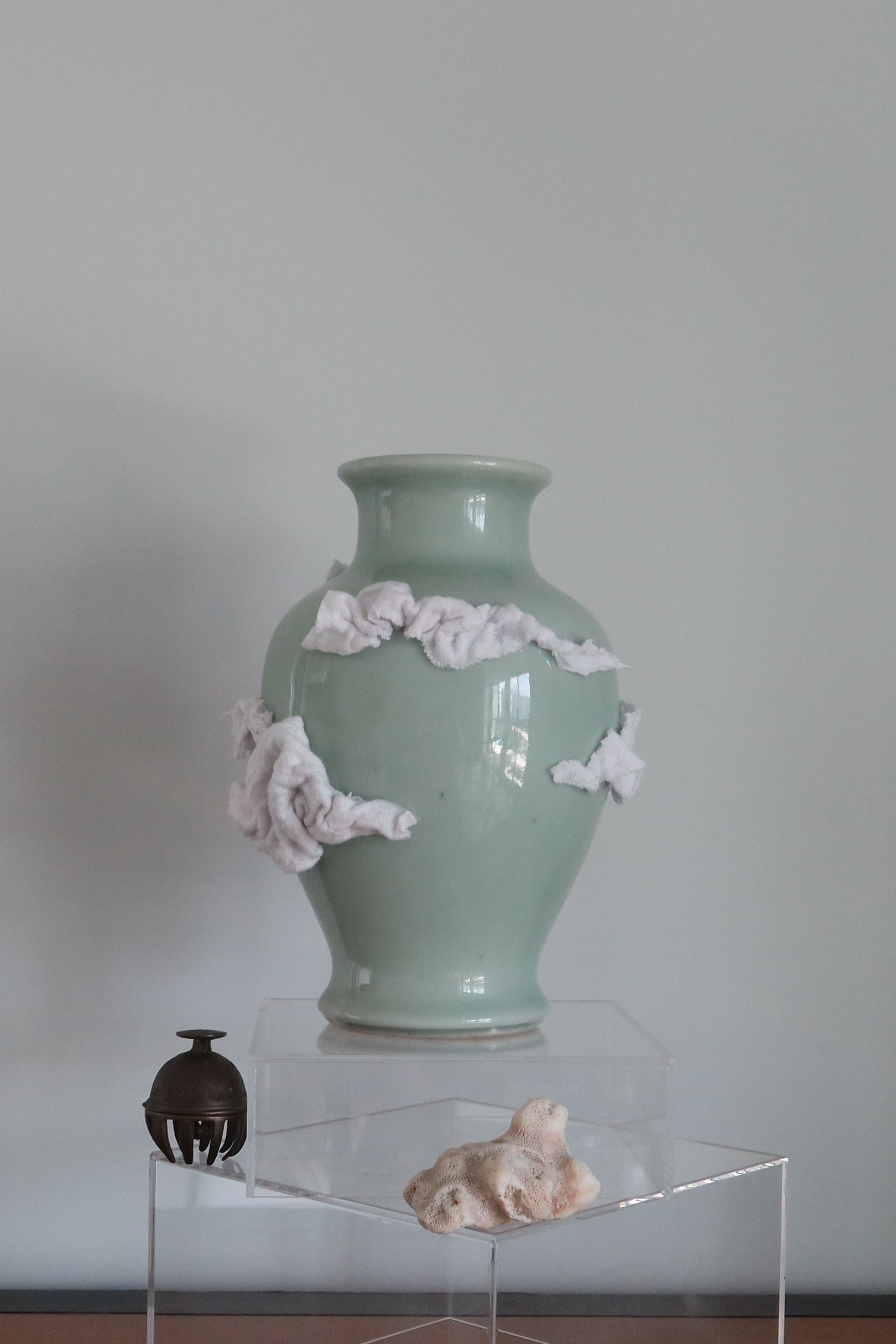 A pale celadon-green ceramic vase with white clouds made of scrap fabric, displayed on a clear acrylic pedestal on a wooden table against a plain light gray wall.