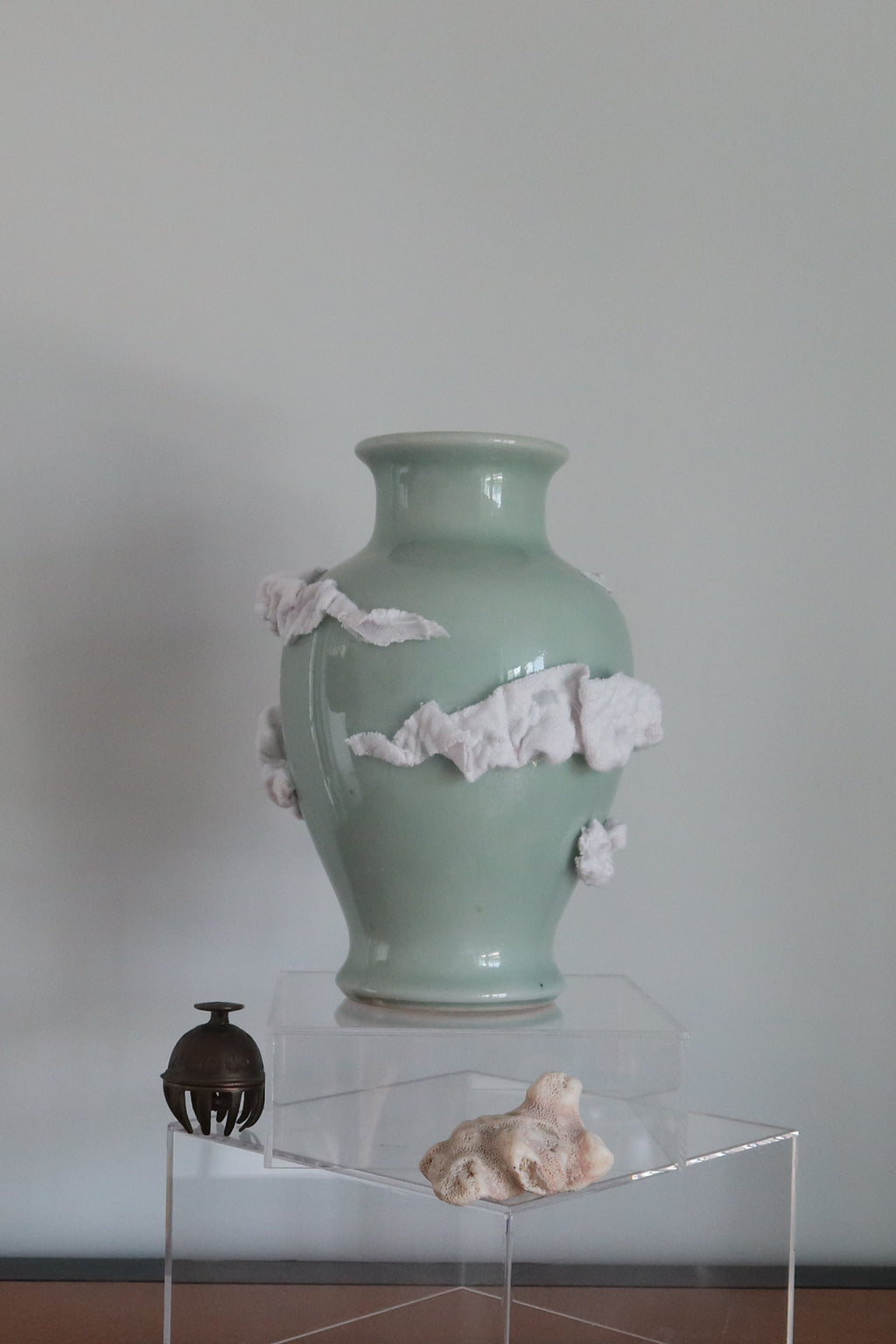 A pale green ceramic vase with white clouds made of fabric as decoration on the vase. It stands on a clear acrylic display block on a wooden table against a plain light gray wall.