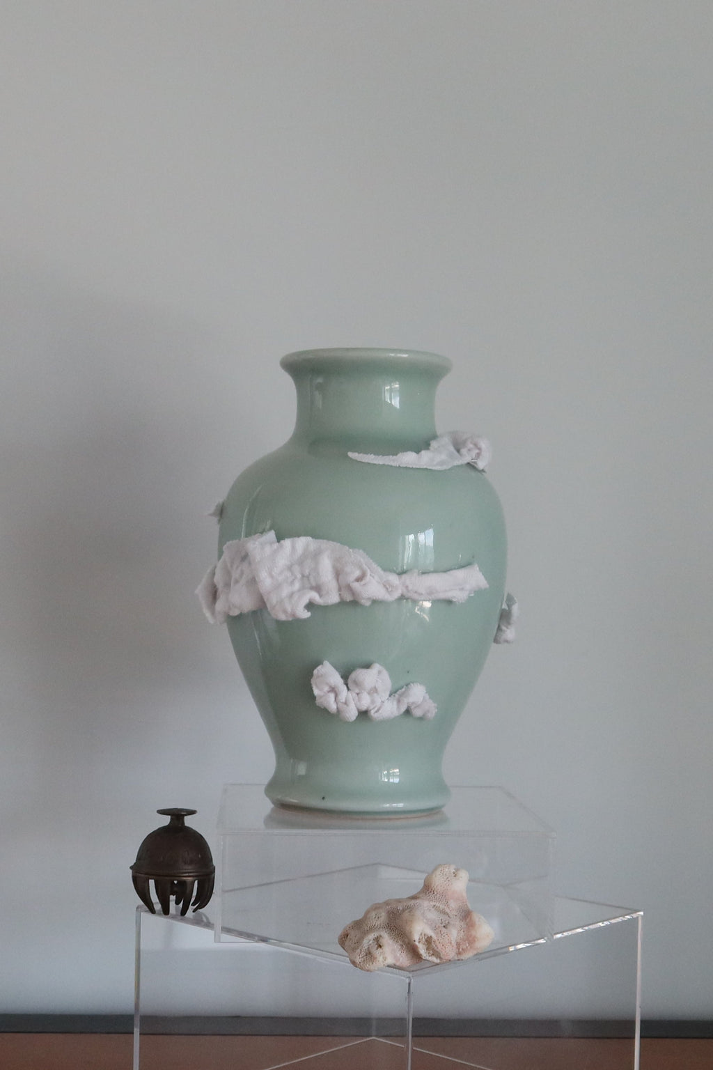 A pale green ceramic vase with white clouds made of fabric as decoration on the vase. It stands on a clear acrylic display block on a wooden table against a plain light gray wall.